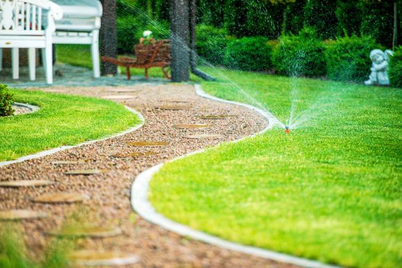 Pelouse verdoyante luxuriante avec arroseur automatique, chemin de pierre et banc blanc dans un cadre de jardin.