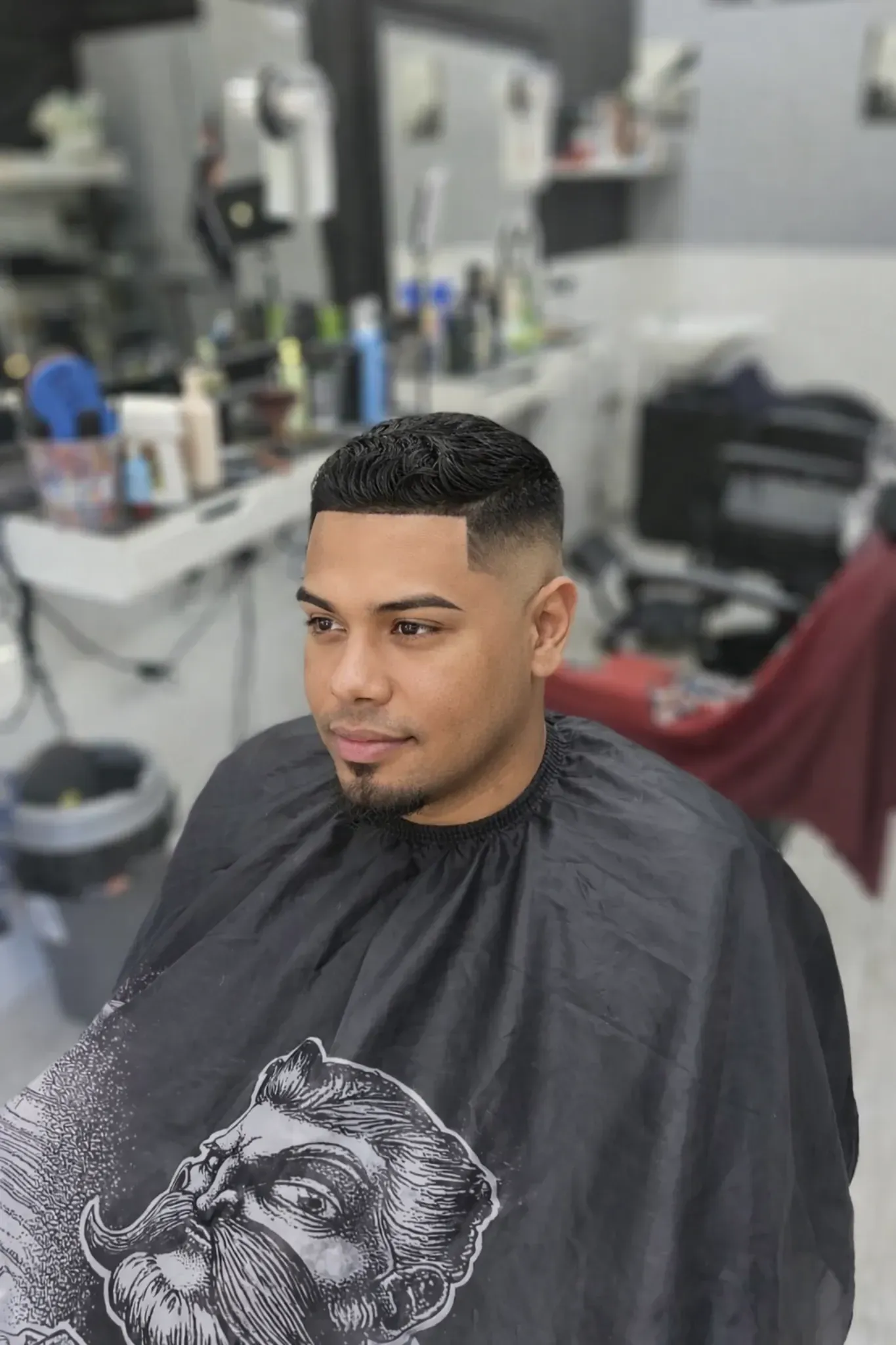Una persona con el pelo corto y descolorido y la barba recortada, sentada con una capa de barbero en una peluquería.