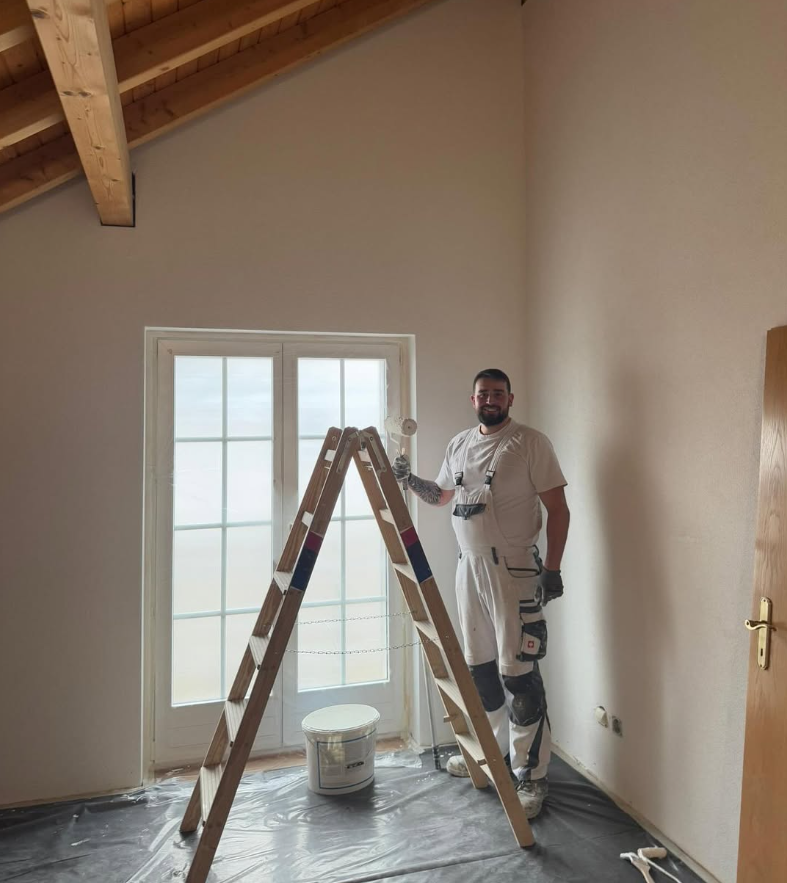 Person, die mit einem Pinsel weiße Zierleisten um ein Fenster streicht.