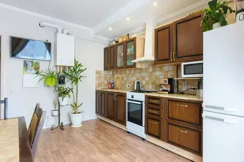 Una cocina con gabinetes marrones, electrodomésticos blancos, piso de madera, plantas y un pequeño televisor.