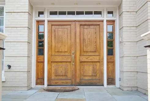 Puertas dobles de madera con ventanales en un edificio de ladrillo claro. Un felpudo de bienvenida reposa sobre los escalones de piedra.