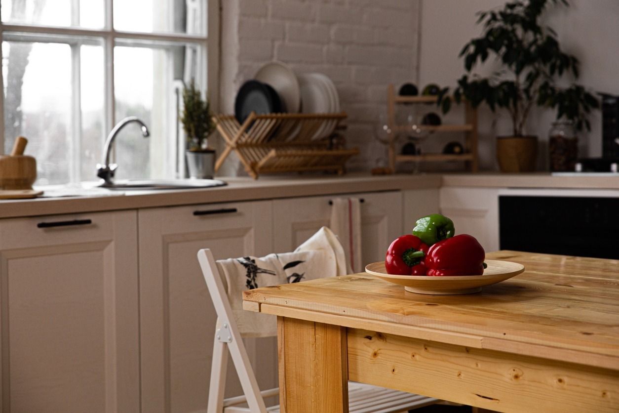 Cocina con mesa de madera, silla y un plato de pimientos rojos y verdes. Fregadero, ventana, platos y planta visibles.
