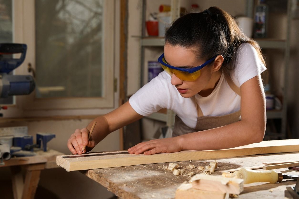 Mujer con gafas de seguridad midiendo madera en un taller, expresión concentrada.