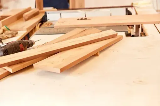 Tablones de madera sobre una sierra de mesa, con un taladro y guantes cerca, para trabajar la madera.