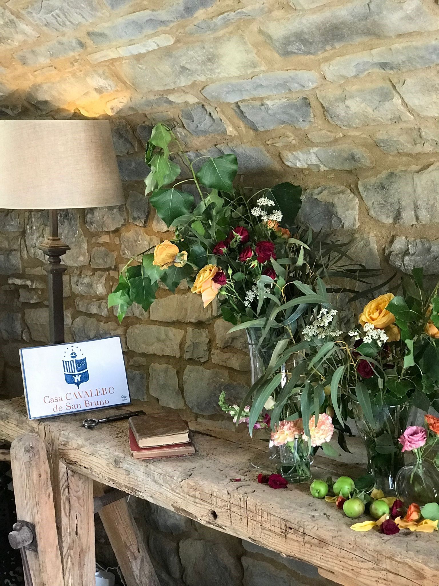 Una mesa de madera con flores y una lámpara encima frente a una pared de piedra.