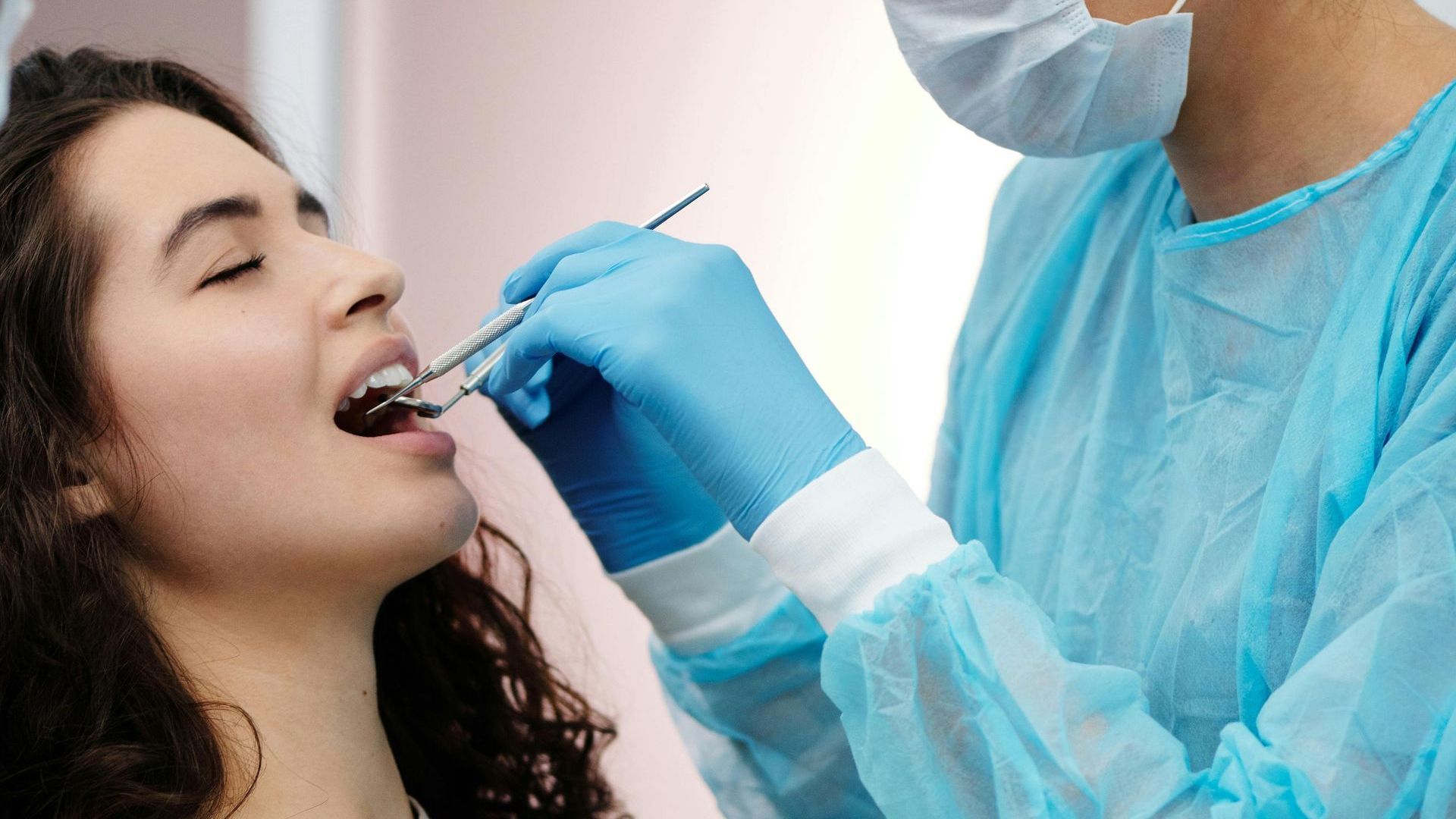 Um dentista examinando os dentes de uma mulher; a mulher está com a boca aberta, e o dentista usa máscara e luvas.