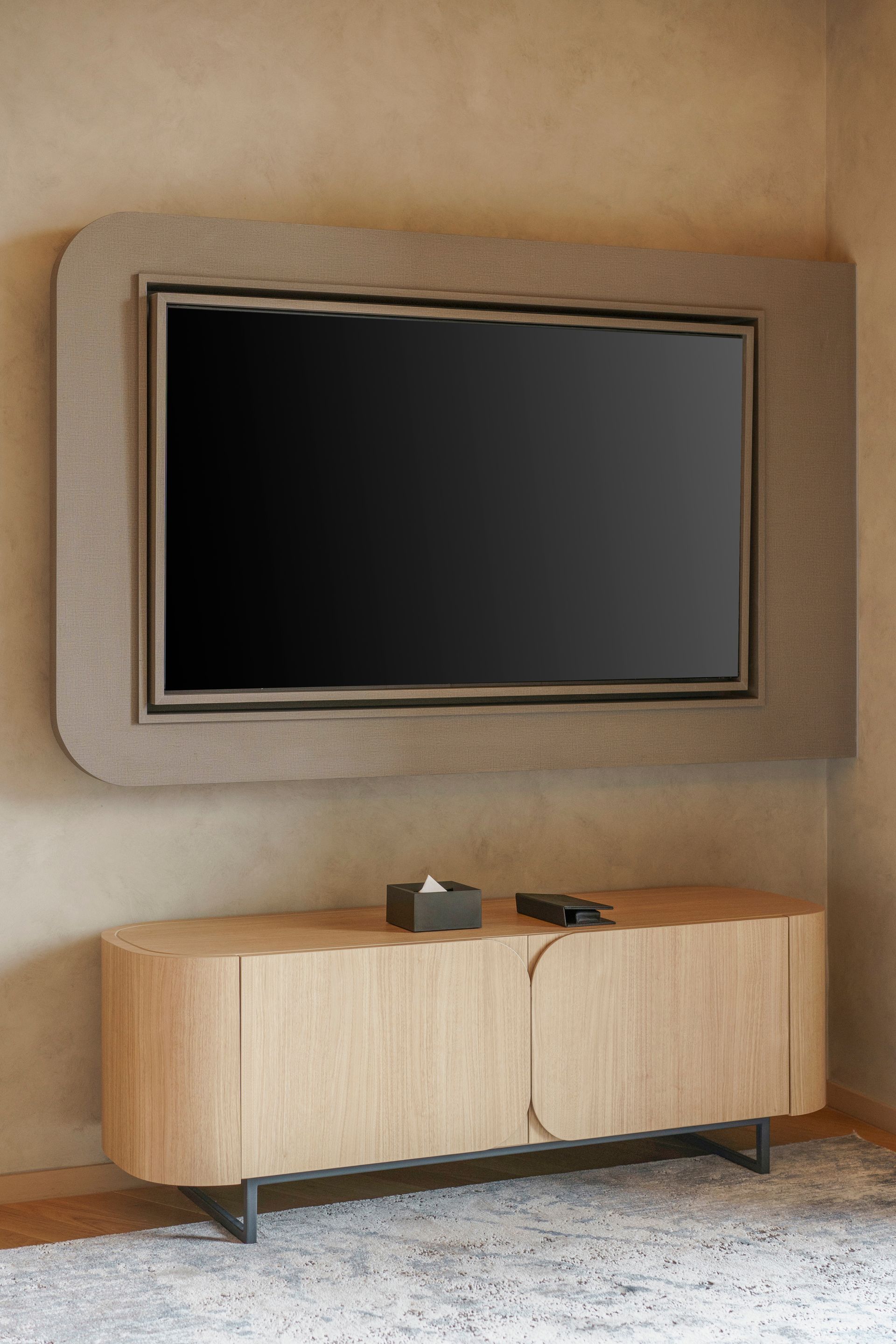 Un téléviseur fixé au mur sur un panneau texturé, au-dessus d'un meuble moderne en bois clair aux bords arrondis.