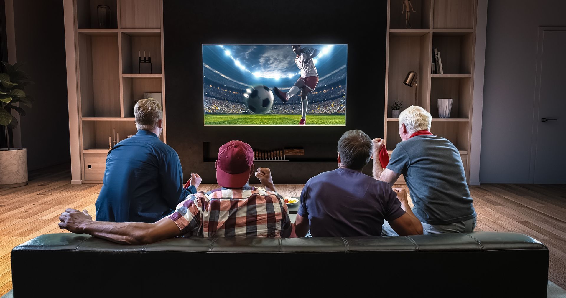 Quatre personnes sont assises sur un canapé dans un salon, regardant un match de football sur un grand écran de télévision.