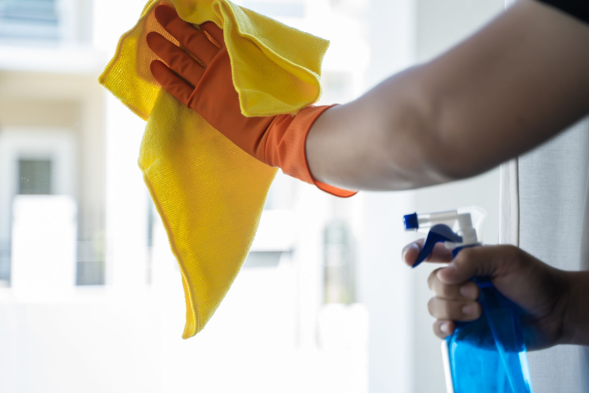 Personne nettoyant une fenêtre avec un chiffon jaune et un vaporisateur bleu.