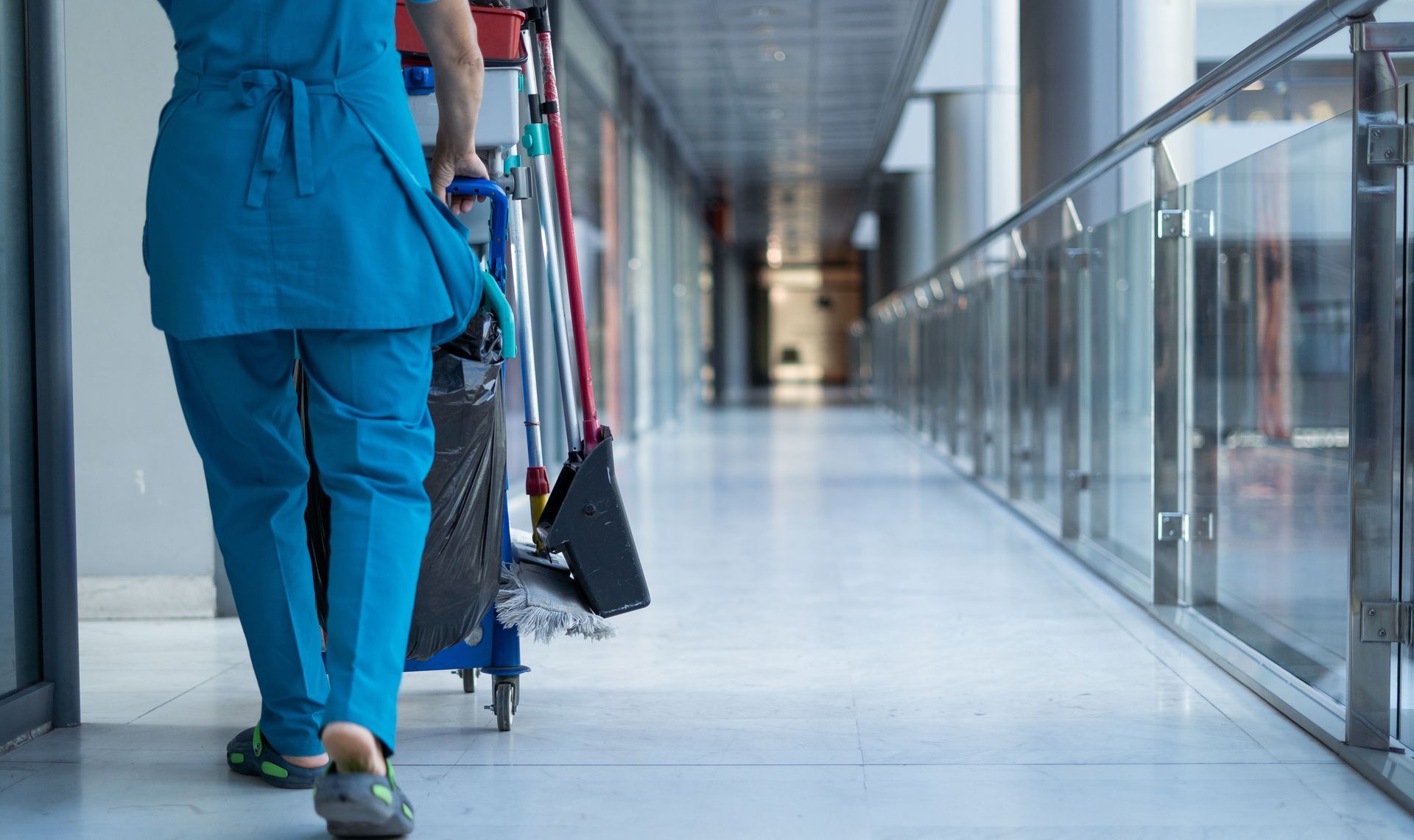 Une personne en tenue bleue pousse un chariot de nettoyage dans un couloir lumineux.