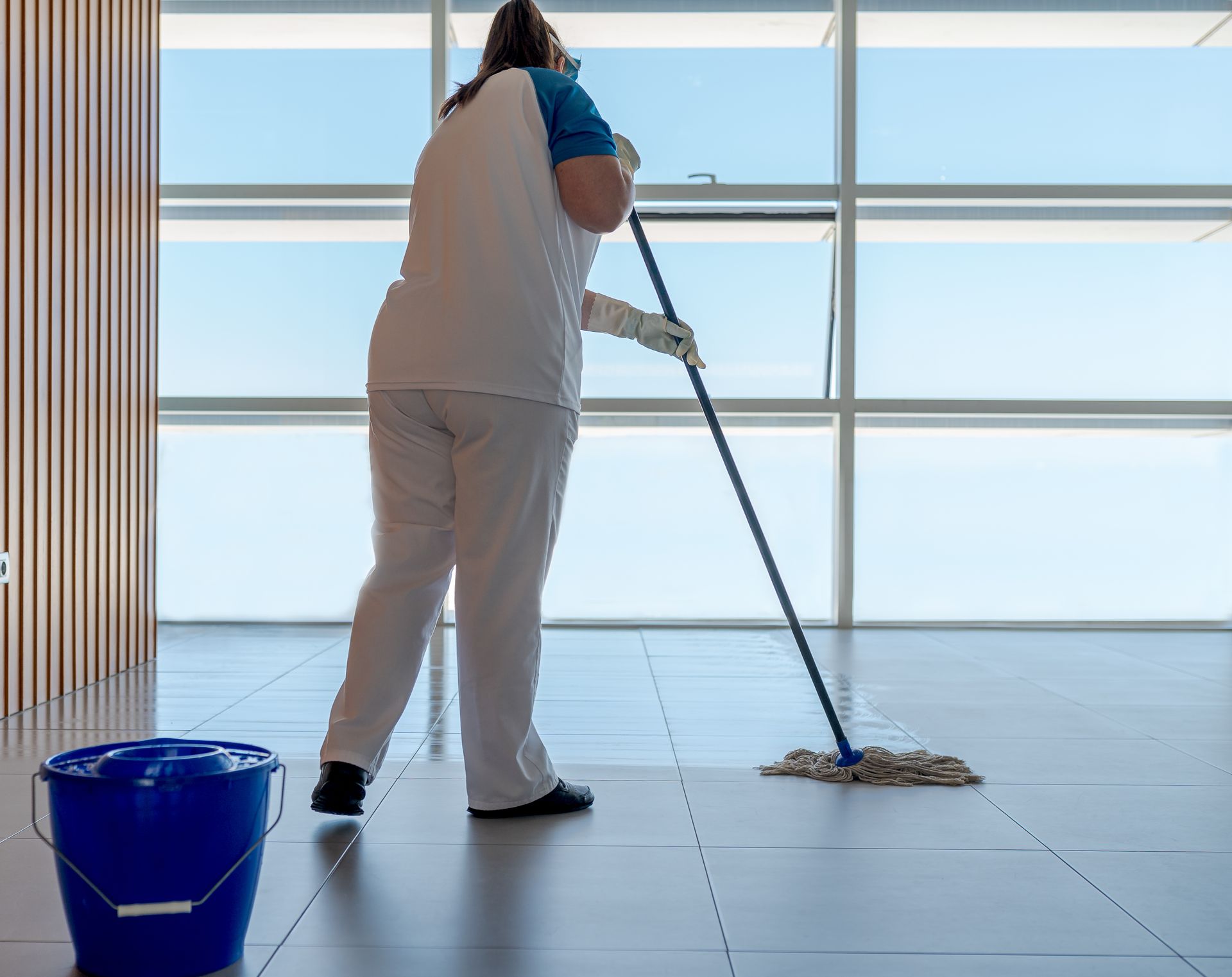 Personne nettoyant un sol carrelé près d'une fenêtre. Seau et serpillière bleus, pantalon, chemise et gants blancs.