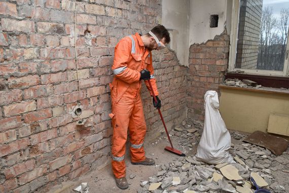 Une personne en combinaison orange balaie les débris dans une pièce éventrée avec des briques apparentes et du plâtre déchiré.