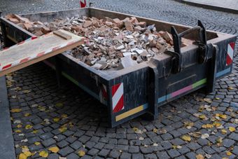 Benne de construction remplie de gravats sur une rue pavée, avec une rampe et des marquages de sécurité.