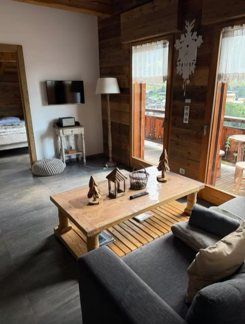 Salon confortable avec boiseries, canapé gris et table basse en bois, s'ouvre sur un balcon.