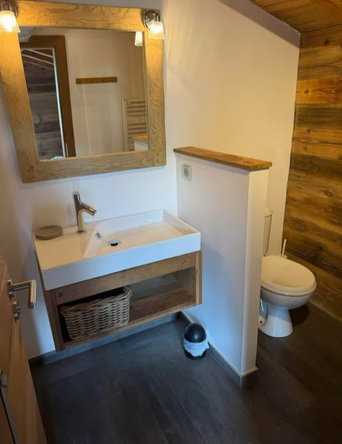 Salle de bain avec lavabo et toilettes blancs, touches de bois et sols foncés.