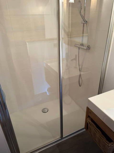 Une petite douche blanche avec une porte coulissante en verre. Des robinetteries chromées et un lavabo blanc sont visibles.