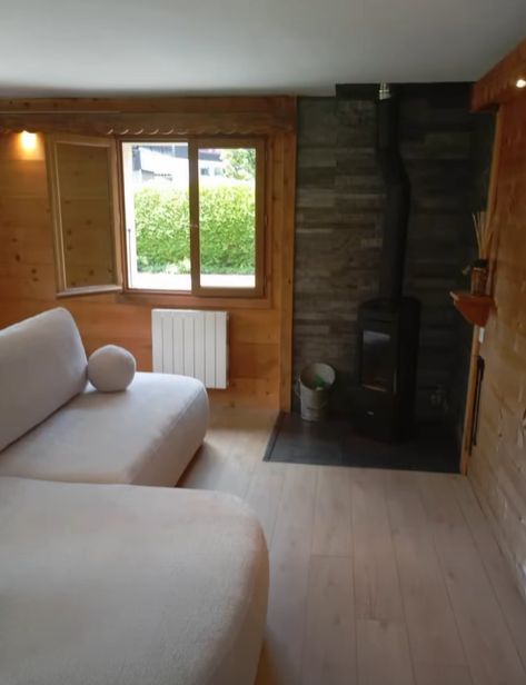 Salon chaleureux avec boiseries, canapé clair, fenêtre et cheminée avec poêle noir.