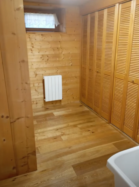 Chambre lambrissée avec portes à lattes de bois, radiateur et petite fenêtre ; parquet et lavabo blanc dans le coin.