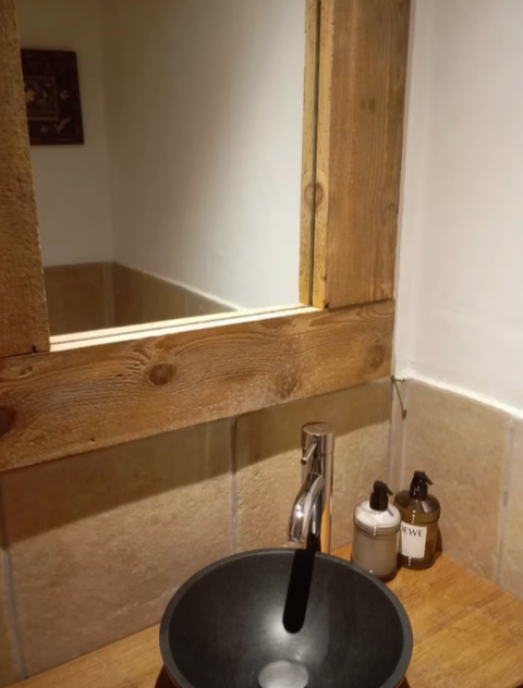 Salle de bain avec miroir à cadre en bois au-dessus d'un lavabo foncé et de robinets, avec distributeurs de savon.