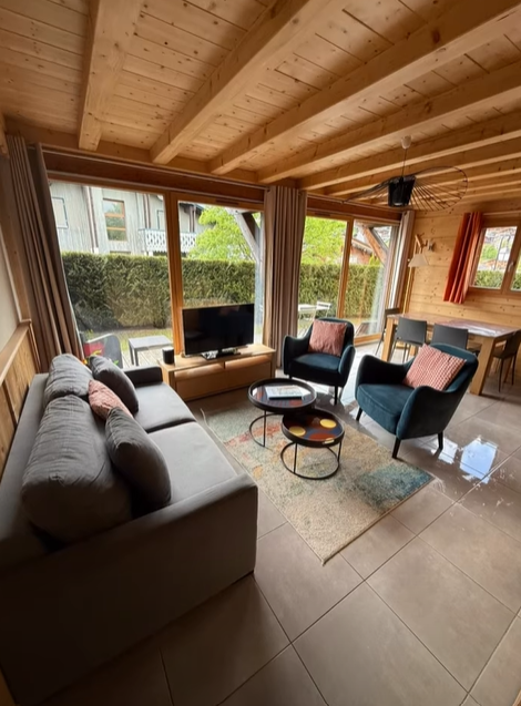 Salon chaleureux avec canapé gris, fauteuils, télévision et table près de grandes fenêtres. Plafond et parquet en bois.