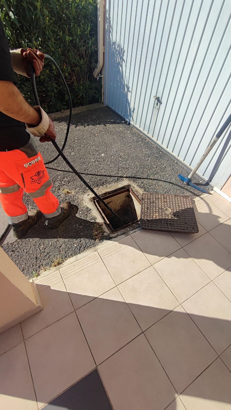 Un ouvrier ouvre un drain dans une terrasse carrelée, à côté d'un mur en tôle ondulée.