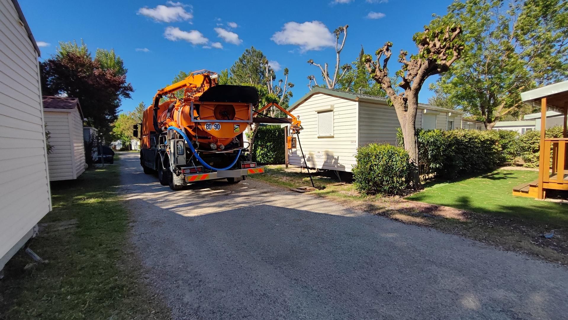 Un camion-citerne orange sur une allée de gravier entre des maisons blanches, sous un ciel bleu arboré.