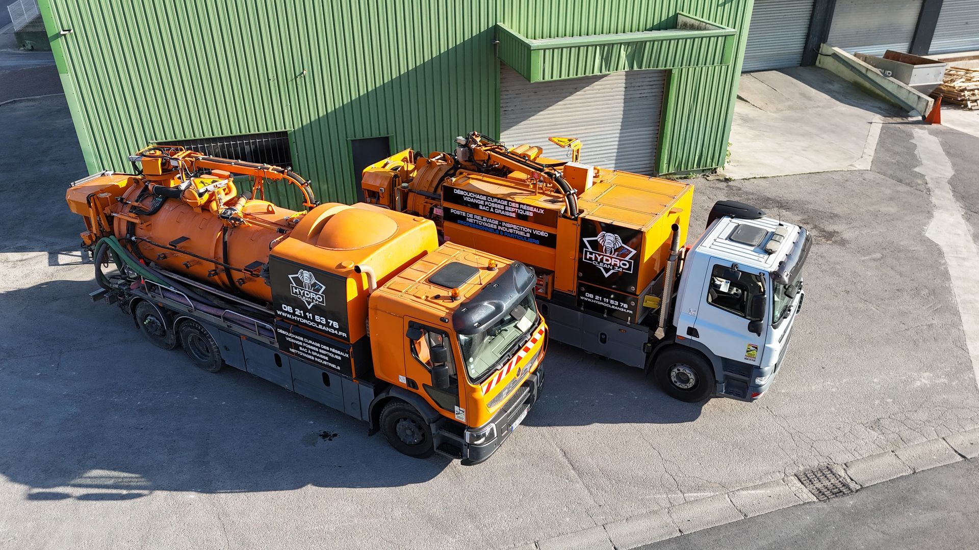 Deux camions utilitaires orange sont garés à côté d'un bâtiment industriel vert sur un terrain ensoleillé.
