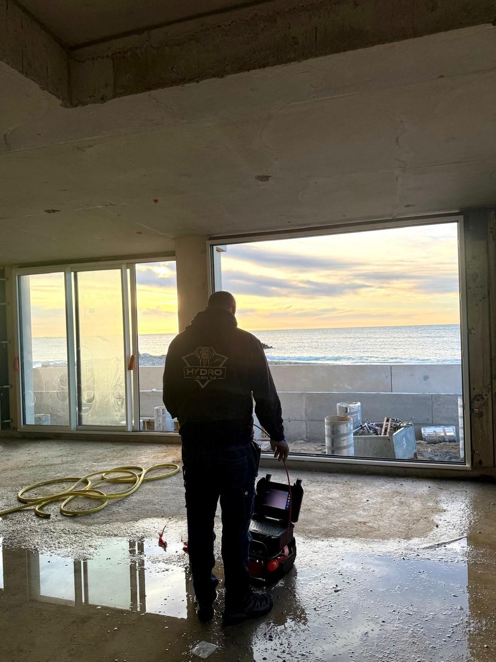 Une personne réalise une inspection vidéo de canalisation dans une pièce vide avec de grandes fenêtres et un sol mouillé au coucher du soleil