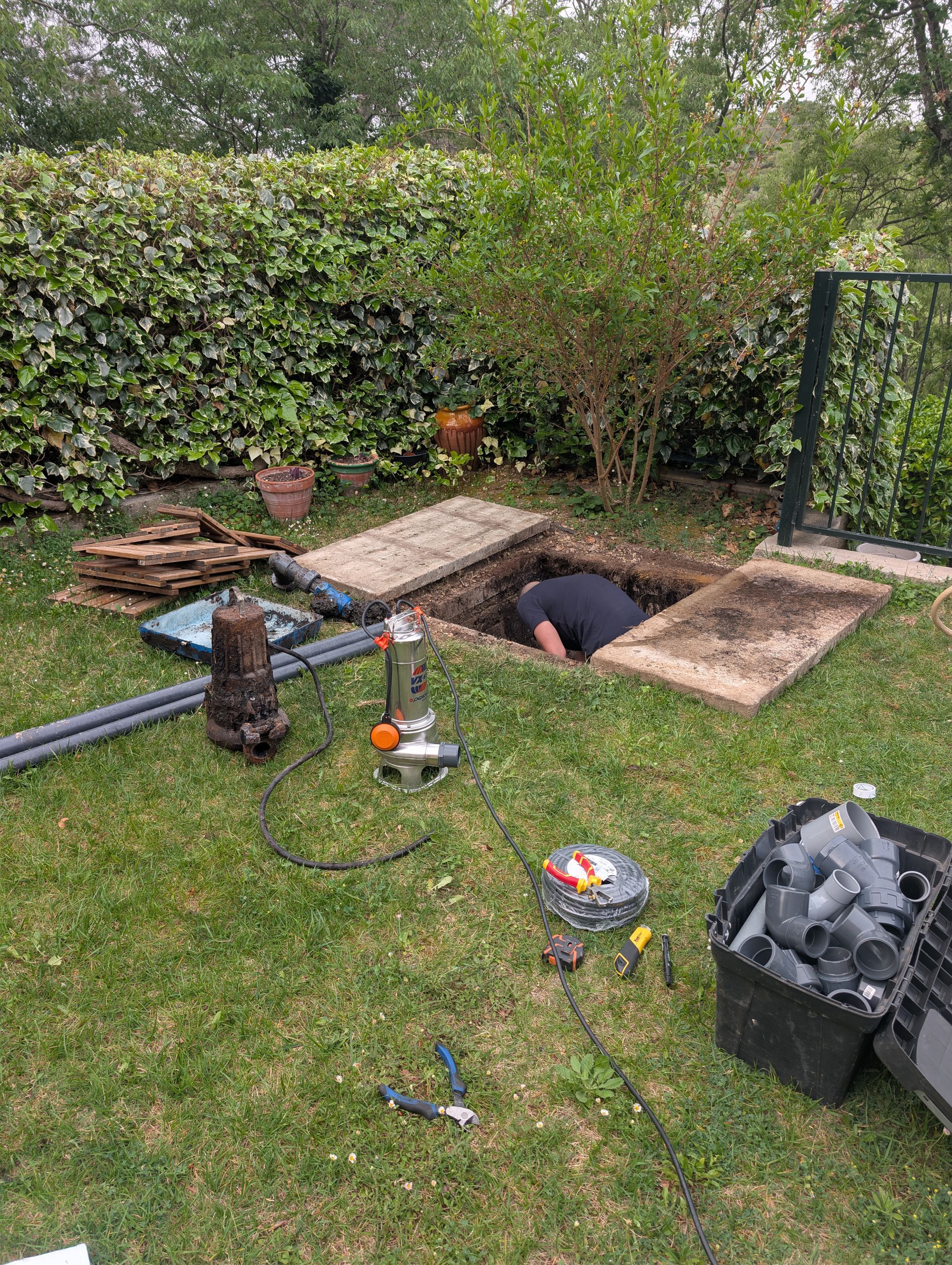 Une personne travaillant dans un jardin, penchée dans un trou rectangulaire à côté de bois et d'outils empilés.