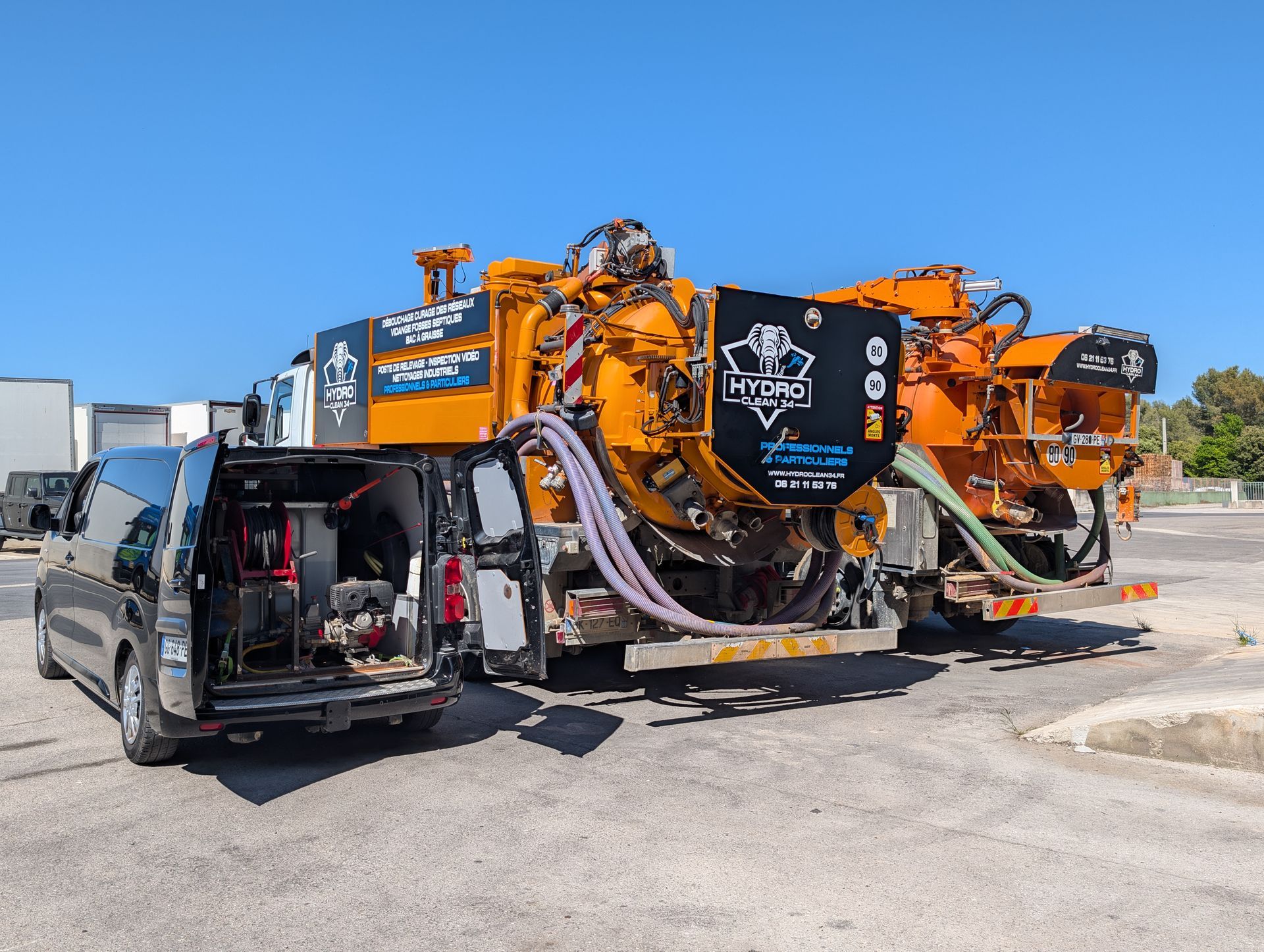 Un camion aspirateur orange à côté d'une camionnette noire sur un parking sous un ciel bleu dégagé