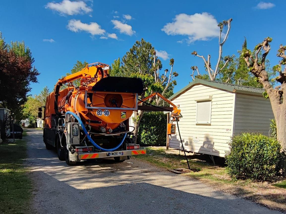 Un camion utilitaire orange avec des équipements surélevés est garé à côté d'un petit hangar blanc sur une allée ensoleillée.