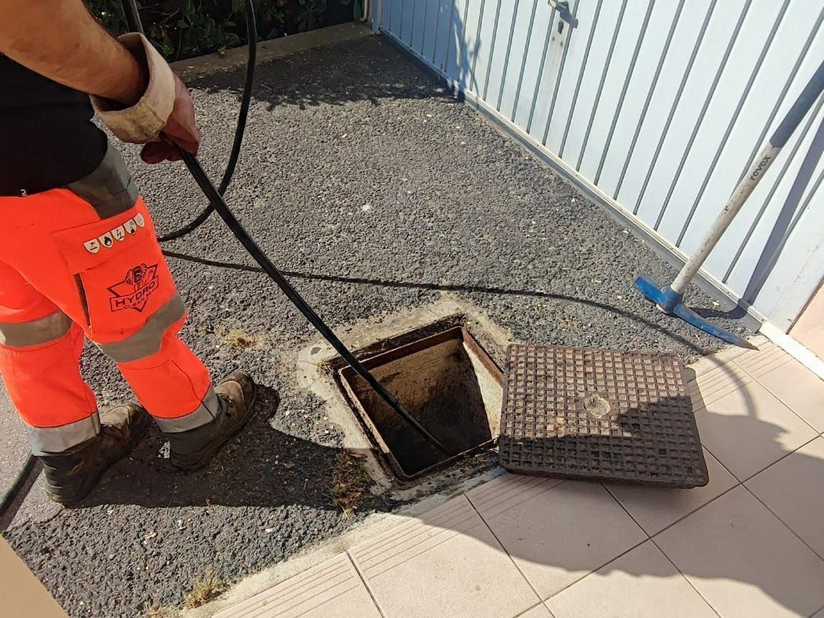 Un ouvrier utilise un outil pour inspecter un regard d'égout ouvert à côté d'un bâtiment blanc.