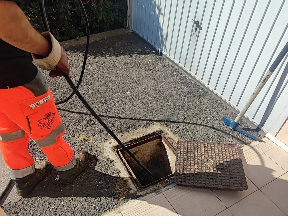Un ouvrier en pantalon de sécurité orange inspecte une trappe de service au sol ouverte, à côté d'une clôture et d'une allée carrelée.