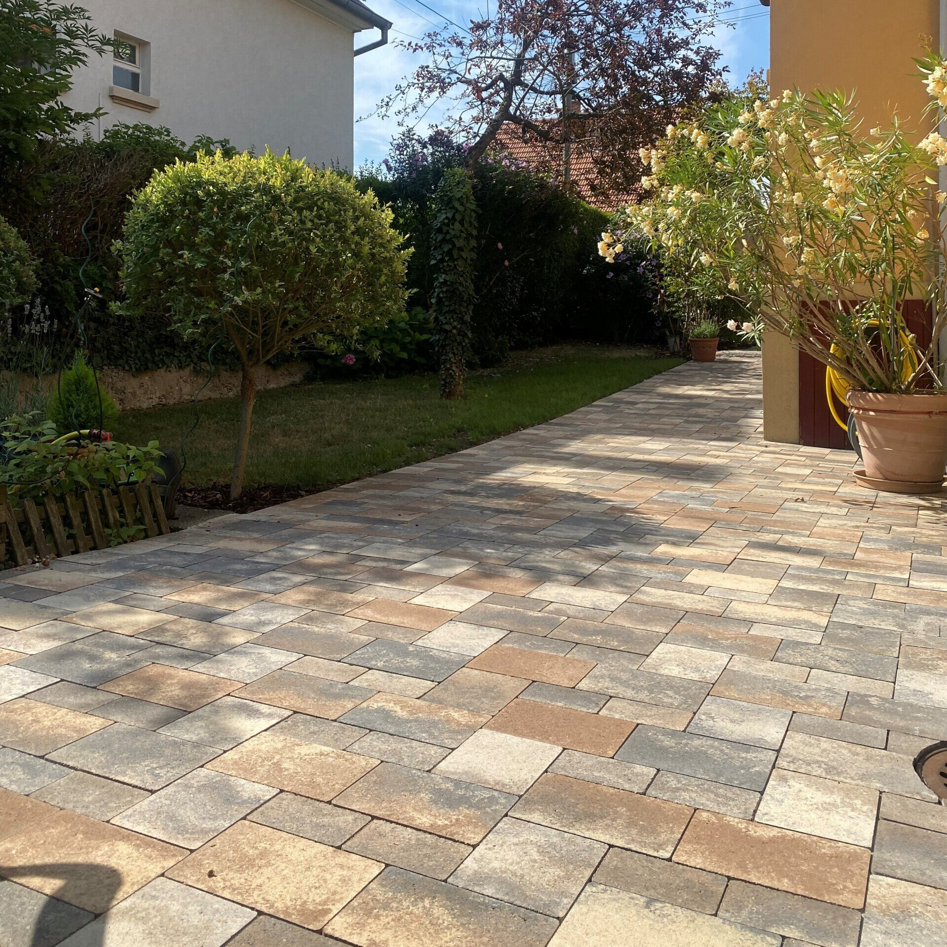 Patio en pierre avec plantes et pelouse menant à une maison, aux tons verts et beiges.