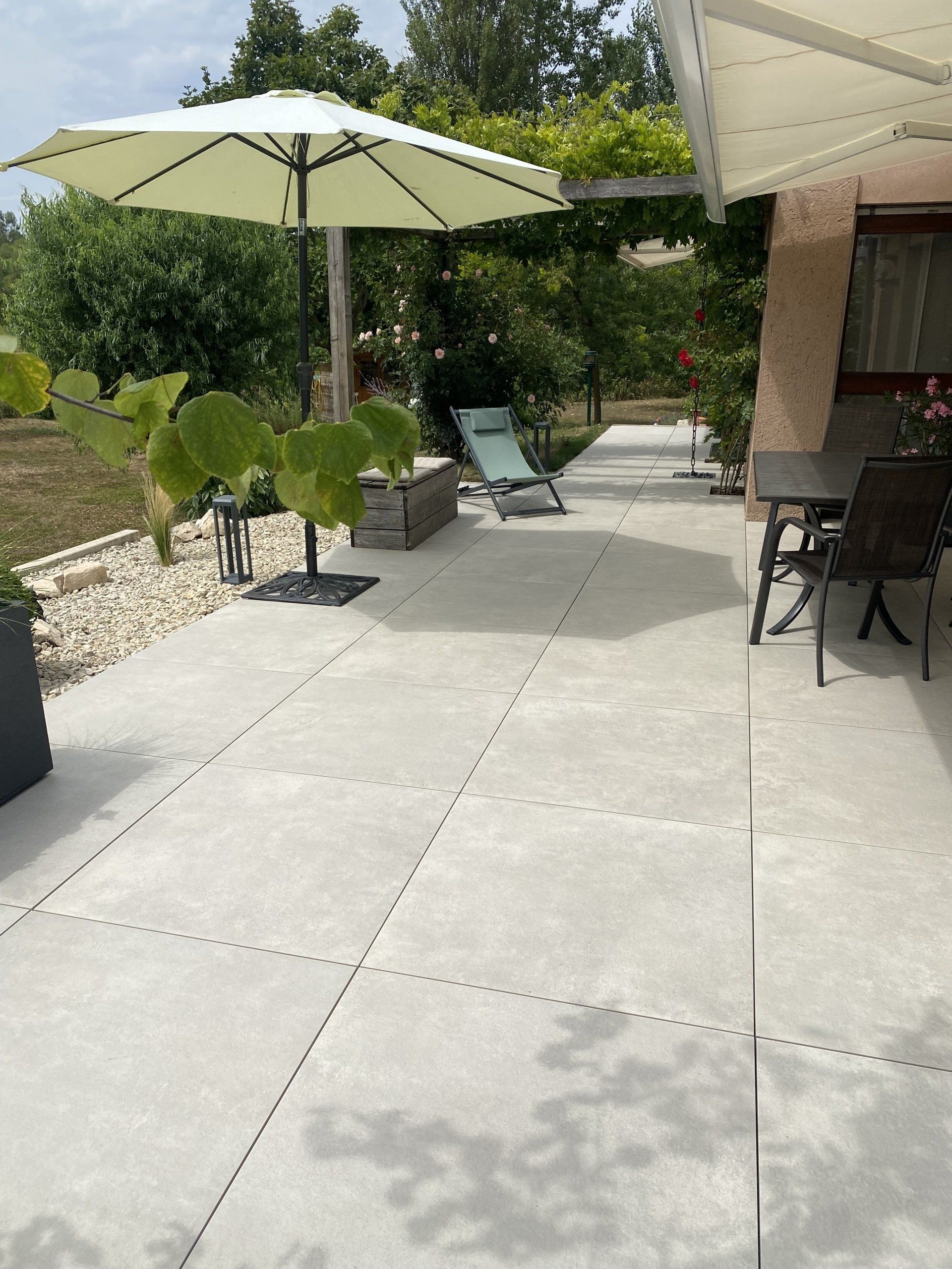 Terrasse avec carrelage gris, chaise longue sous parasol et table à manger extérieure. Arbres en arrière-plan.