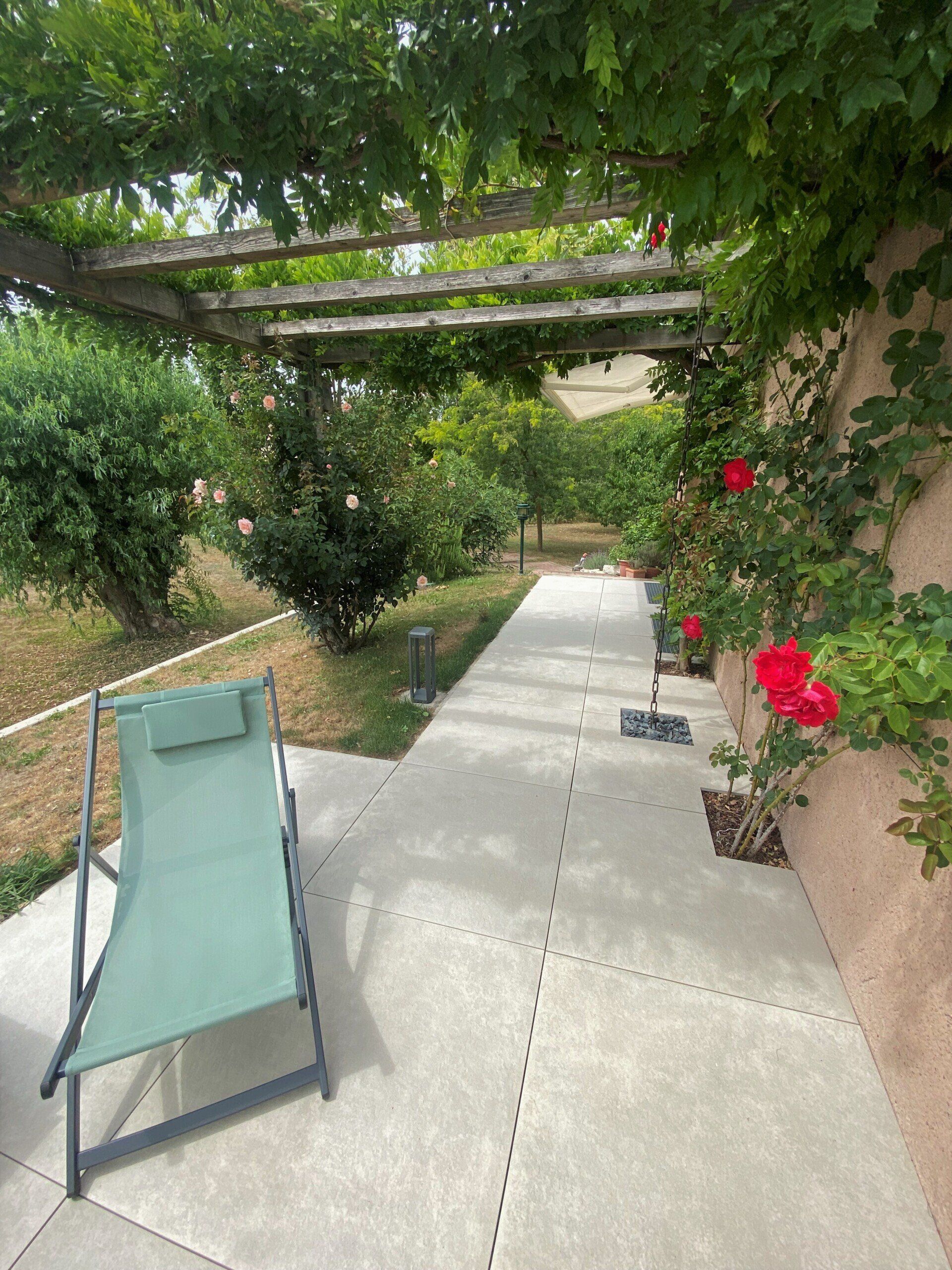 Chaise longue sur une terrasse, surplombée par une pergola recouverte de vigne. Des roses et de la verdure poussent à proximité.