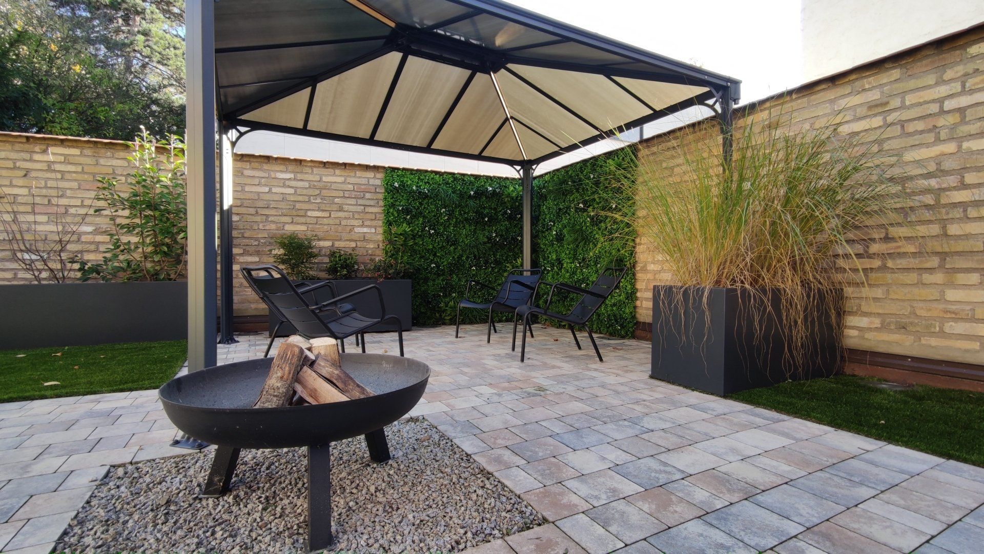 Patio avec foyer, sièges sous gazebo, entouré d'un mur de briques, de verdure et de pavés.