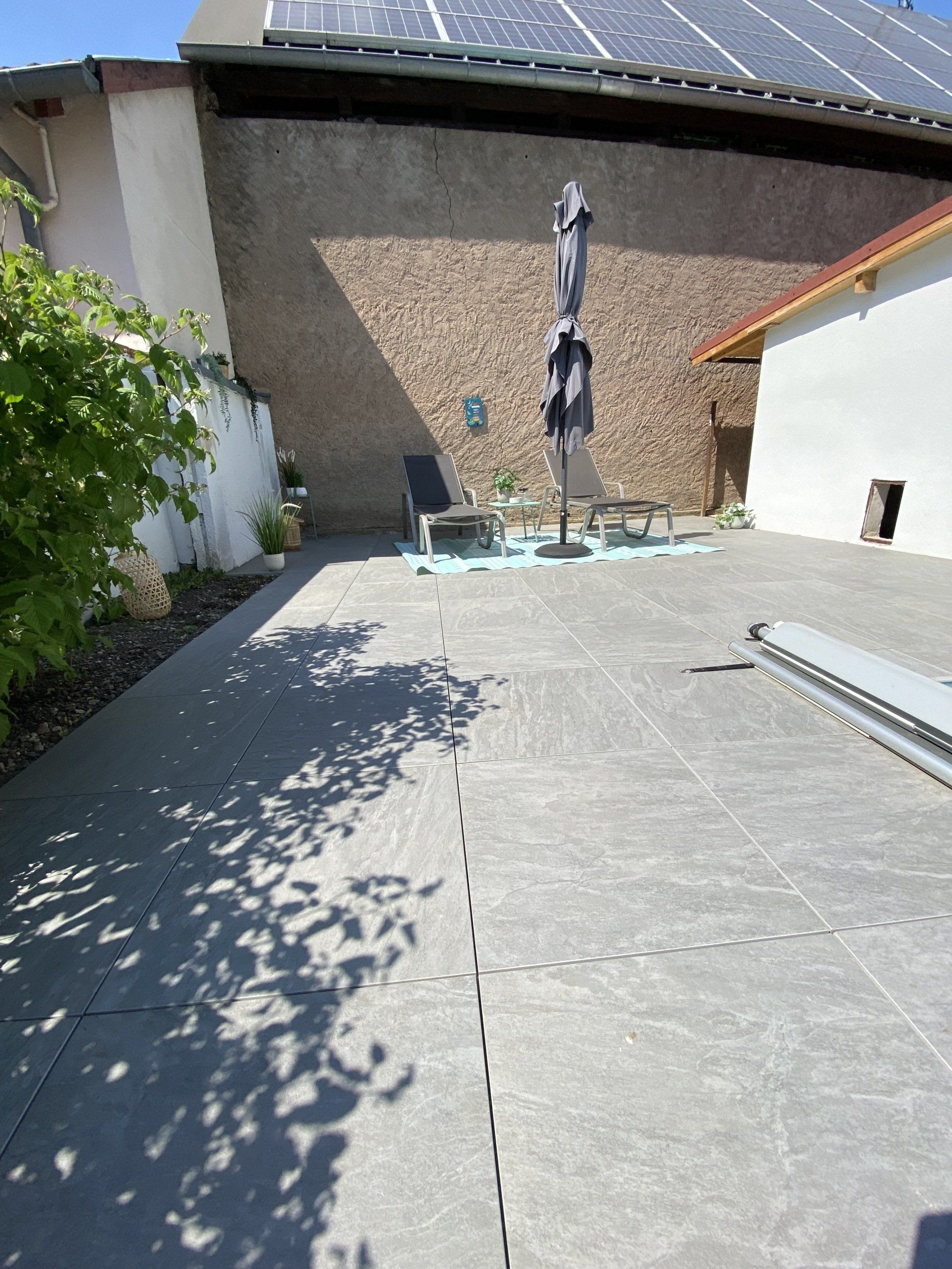 Patio avec carrelage gris, mobilier d'extérieur et parasol, adjacent à des bâtiments avec panneaux solaires.