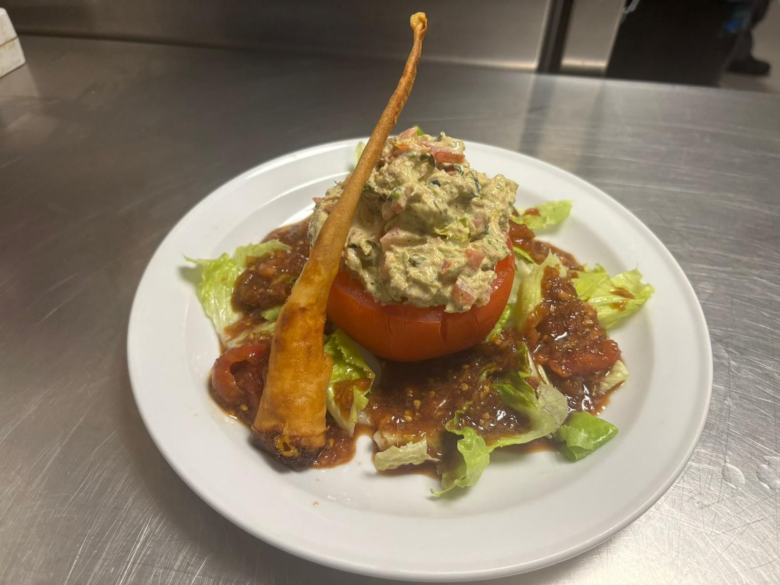 Un tomate relleno de relleno verde reposa sobre lechuga y salsa en un plato blanco; sobresale un palito de masa.