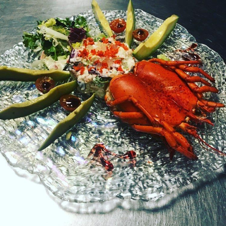 Plato de langosta en un plato transparente con textura. Incluye langosta, ensalada, rodajas de aguacate y tomates cherry.