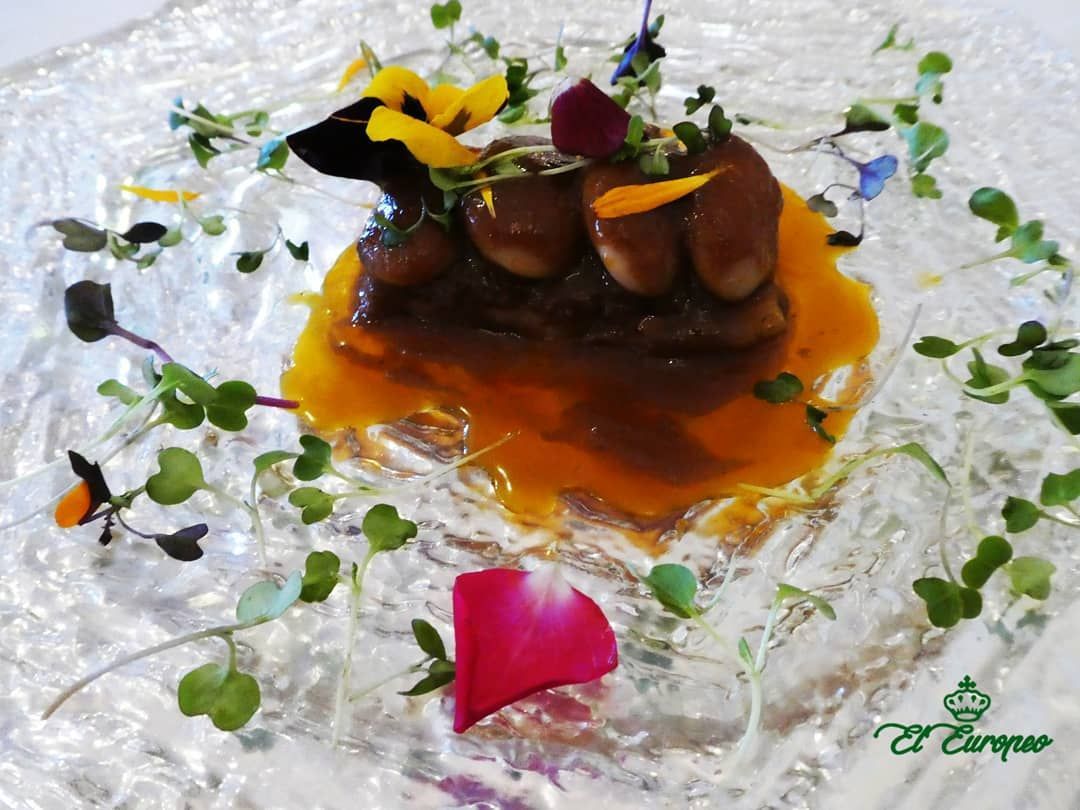 Plato gourmet con carne glaseada, salsa de naranja y flores comestibles en un plato de vidrio decorativo.