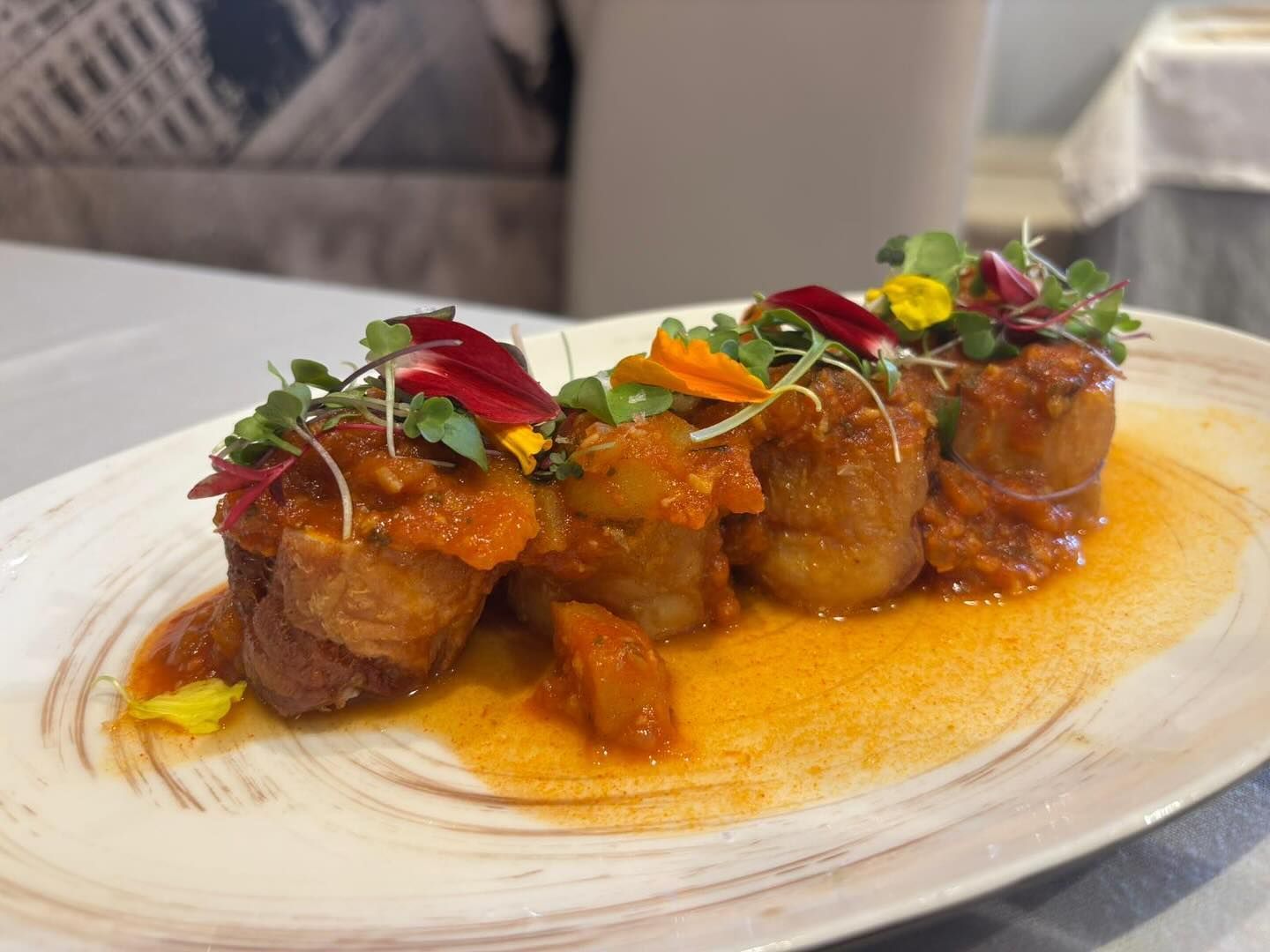 Cuatro rollitos de patata fritos en salsa de tomate, adornados con microvegetales y flores comestibles en un plato estampado.
