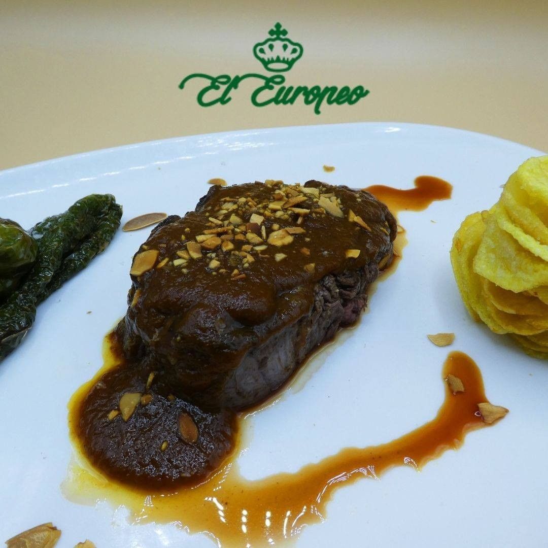 Plato de ternera con salsa, cobertura de almendras, pimientos y patatas fritas en un plato blanco, con el logotipo de fondo.