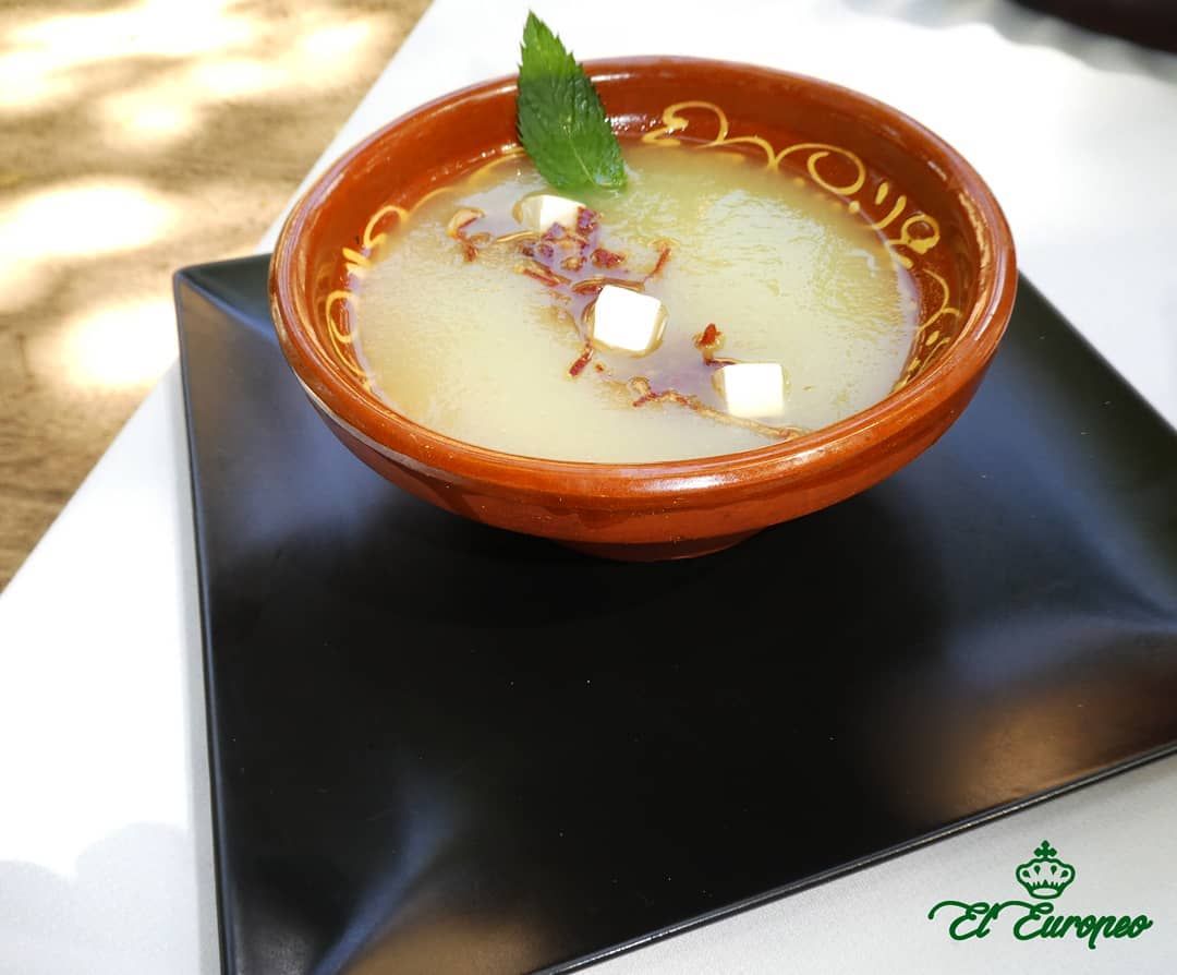 Tazón de sopa fría adornado con menta y cubitos en un plato negro.
