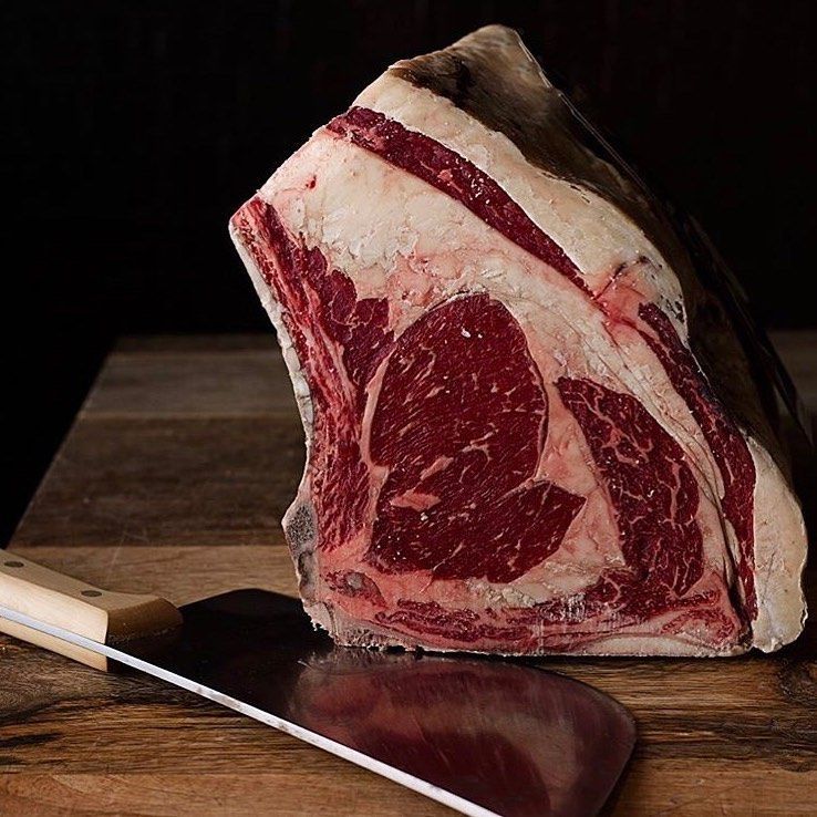 Filete grande de costilla crudo con hueso, colocado sobre una superficie de madera al lado de un cuchillo de carnicero.