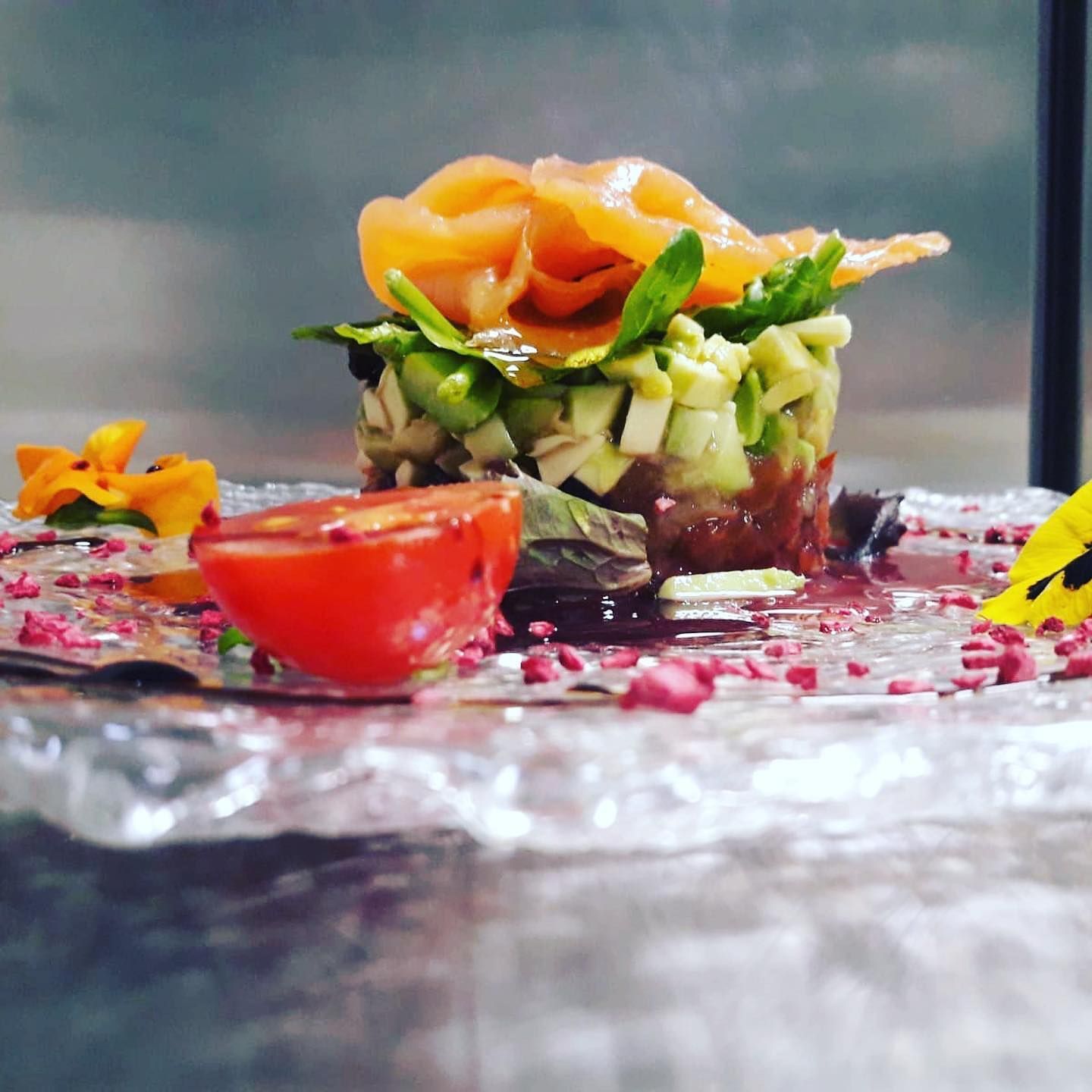 Plato de comida gourmet con torre de salmón, tomate y flores sobre un plato de vidrio transparente.