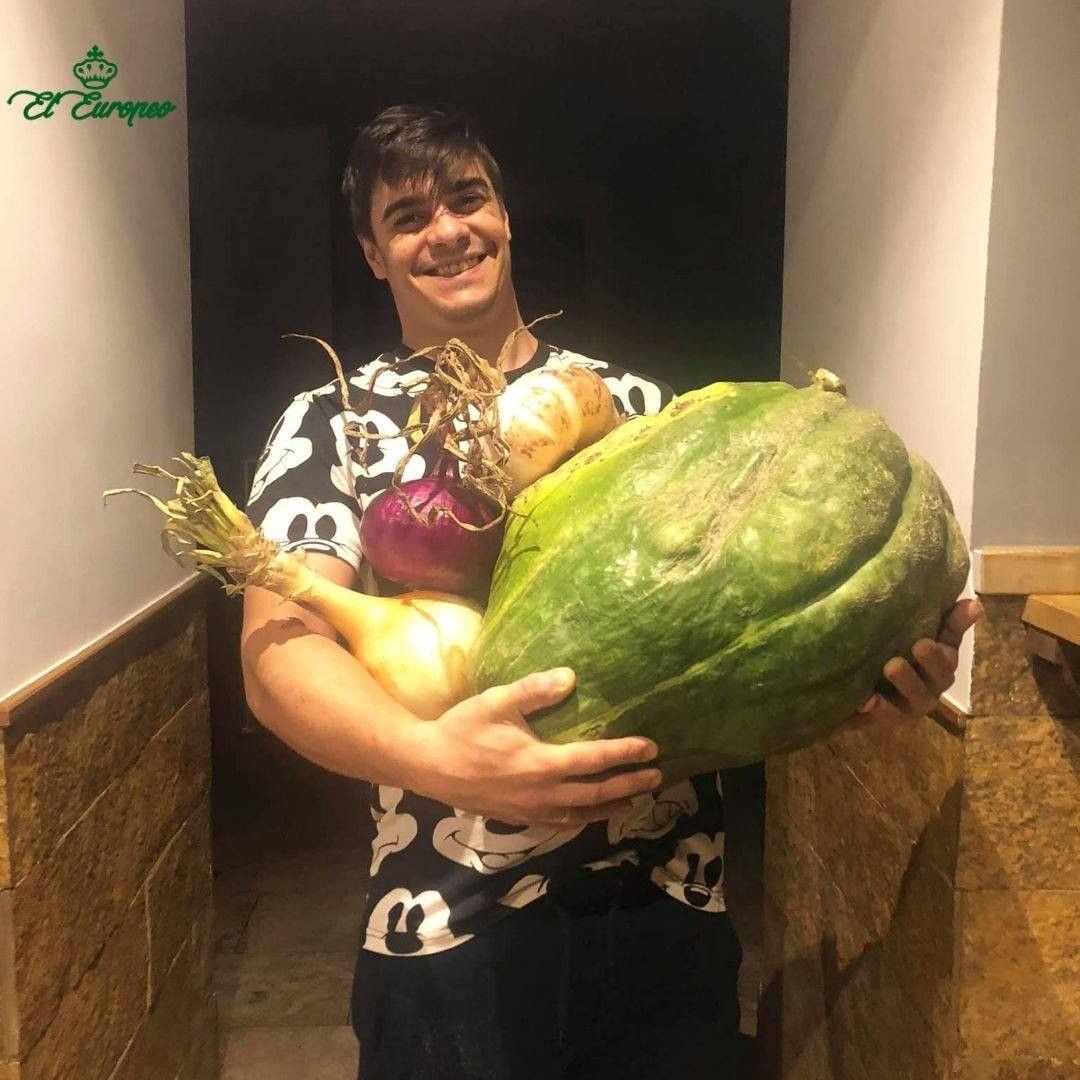 Hombre sosteniendo una calabaza grande y cebollas; sonriendo, de pie en el interior.
