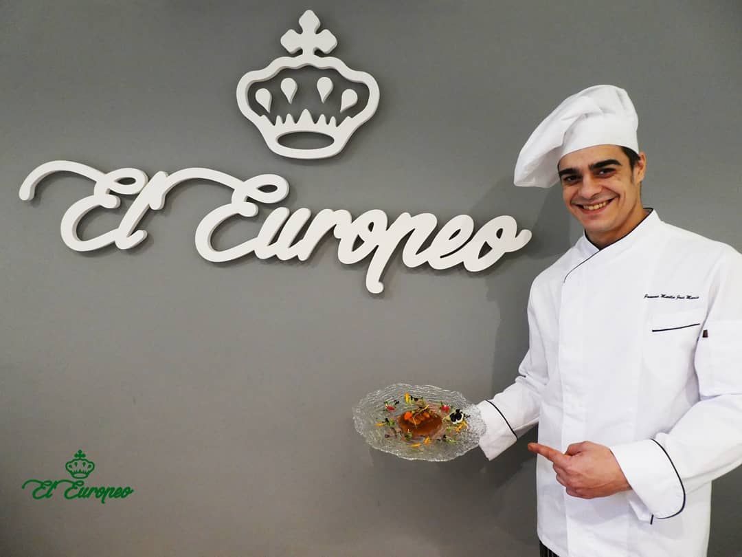 Chef sosteniendo un plato, sonriendo, frente al logo de