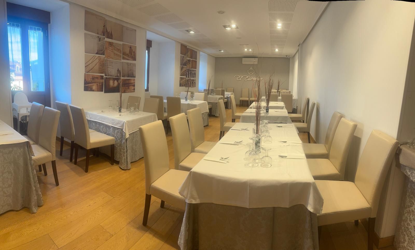 Comedor de restaurante con varias mesas rectangulares preparadas para el servicio. Sillas color crema, manteles beige y suelos de madera clara.