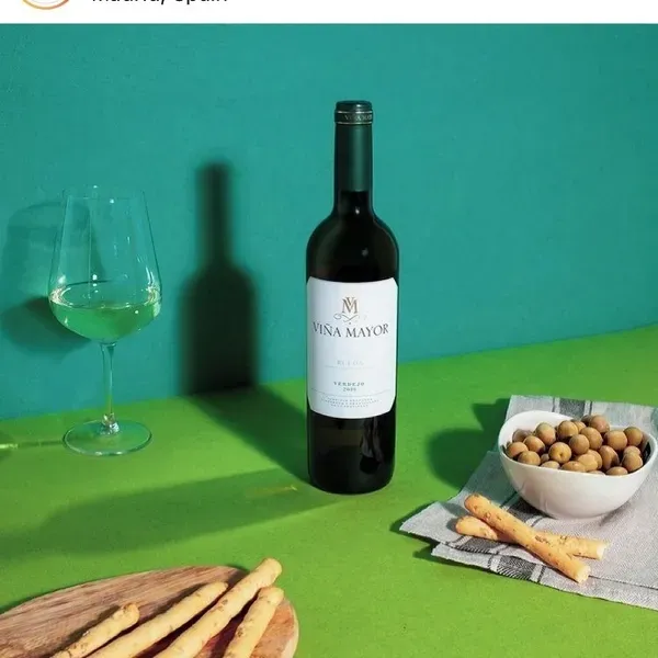 Botella de vino Viña Mayor con copa de vino, snacks sobre superficie verde.